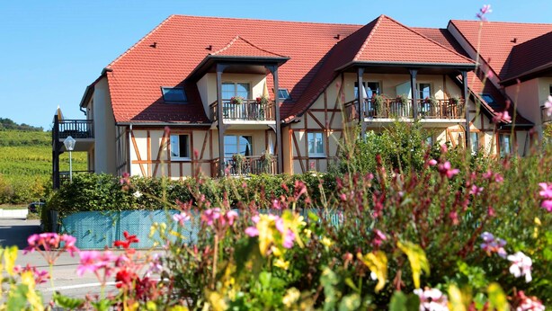 Ein mehrstöckiges Gebäude mit rotem Ziegeldach, Balkonen mit Geländern und einem Gartenbereich im Vordergrund mit verschiedenen Pflanzen und Blumen.