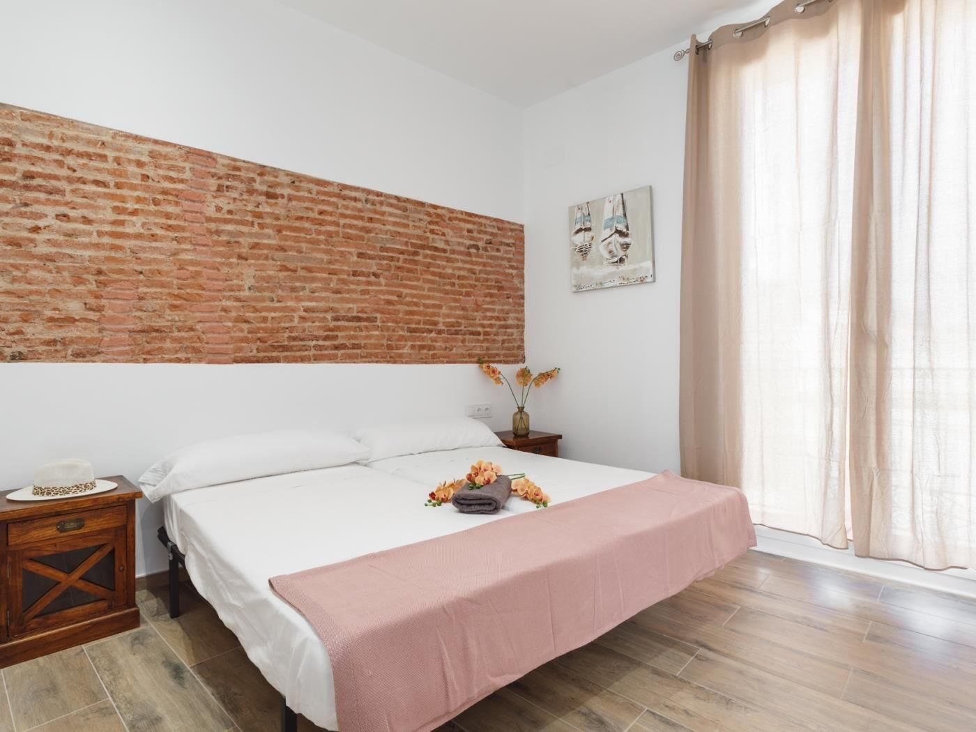 A bedroom with a brick wall, a bed with a pink bedspread, a nightstand, a vase with flowers, and a window with curtains.