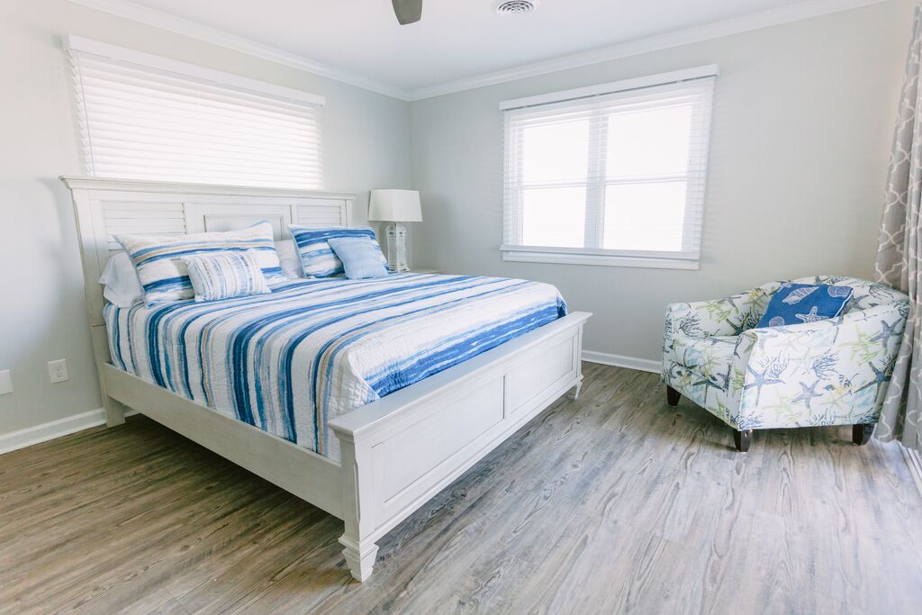 A bedroom with a white bed, striped bedding, a nightstand with a lamp, a chair with a floral pattern, and a window with blinds.