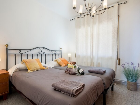 A double bed with a metal headboard, two bedside tables, a chandelier, a window with curtains, and a potted plant.
