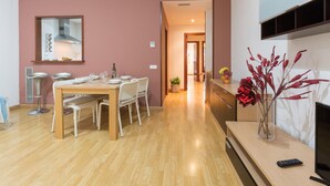 Una sala de estar moderna con una mesa de comedor de madera dispuesta para cuatro personas, un televisor sobre un mueble de madera y un jarrón con ramas rojas.