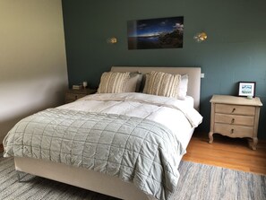 A bedroom with a bed, bedside tables, a nightstand, a framed picture, and a wall-mounted light fixture.