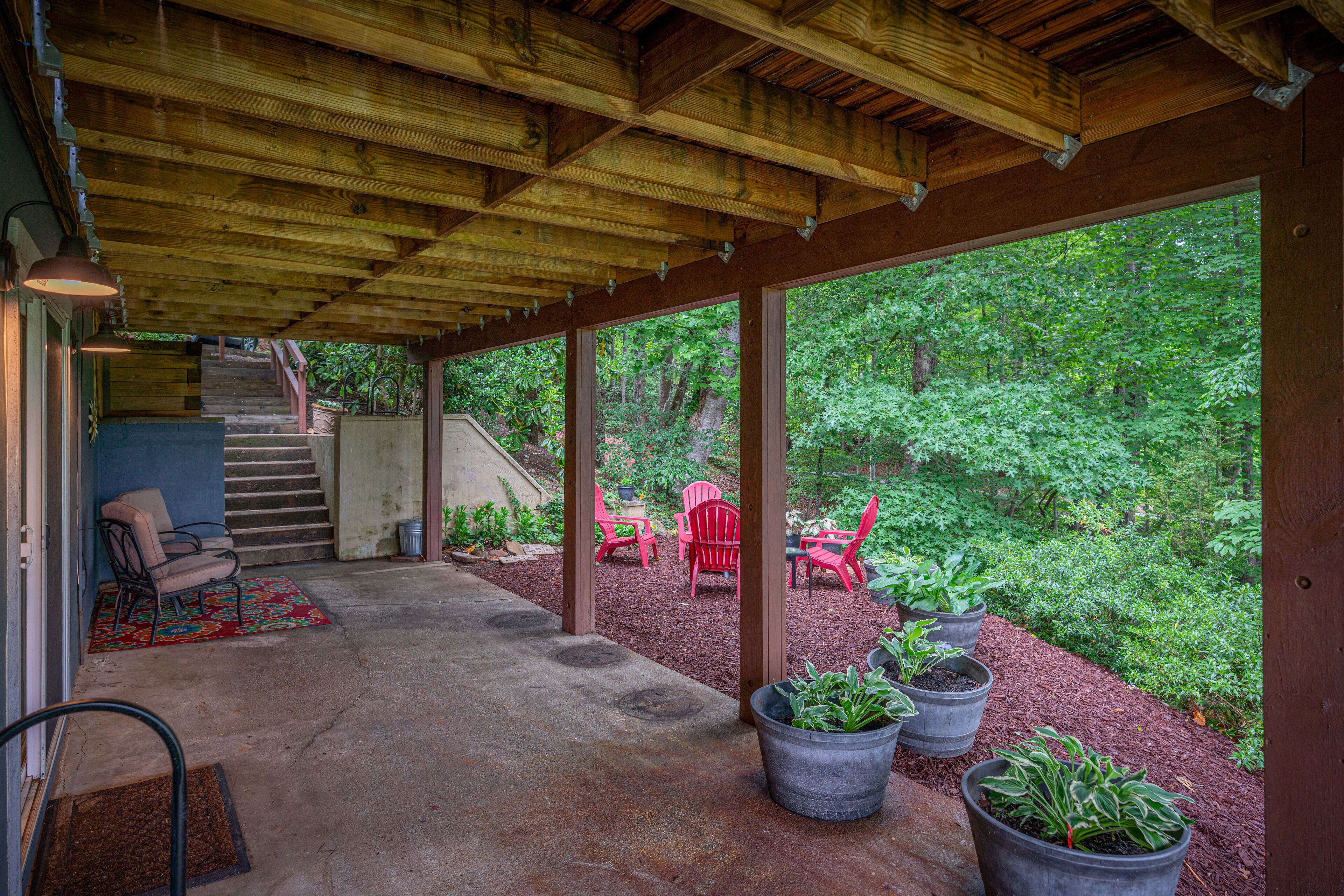 A covered patio with a view of a forest, outdoor seating, and a staircase leading to an upper level.