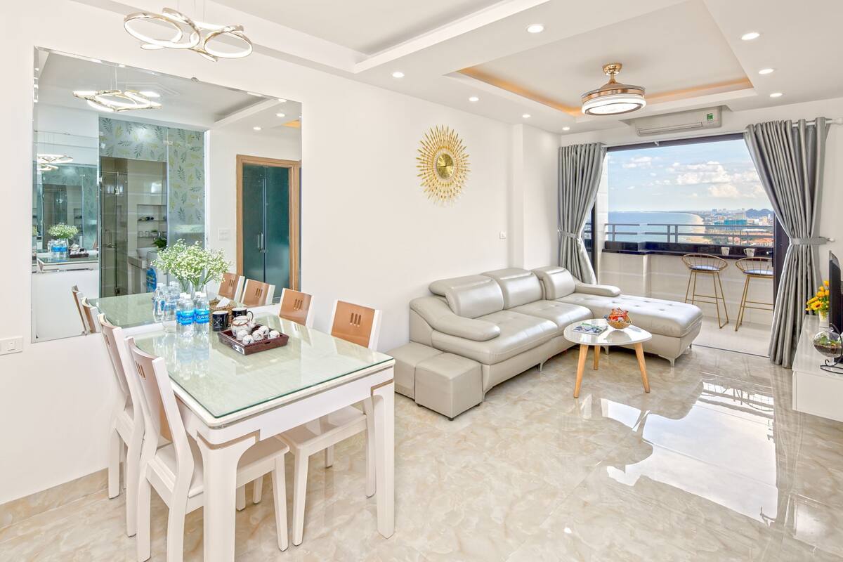 A modern living room with a glass dining table, a sofa, and a view of the sea.