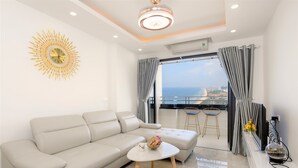 A modern living room with a sofa, a small table, a decorative wall piece, and a view of the sea through large windows. - Muong Thanh Apartment (Da Nang)