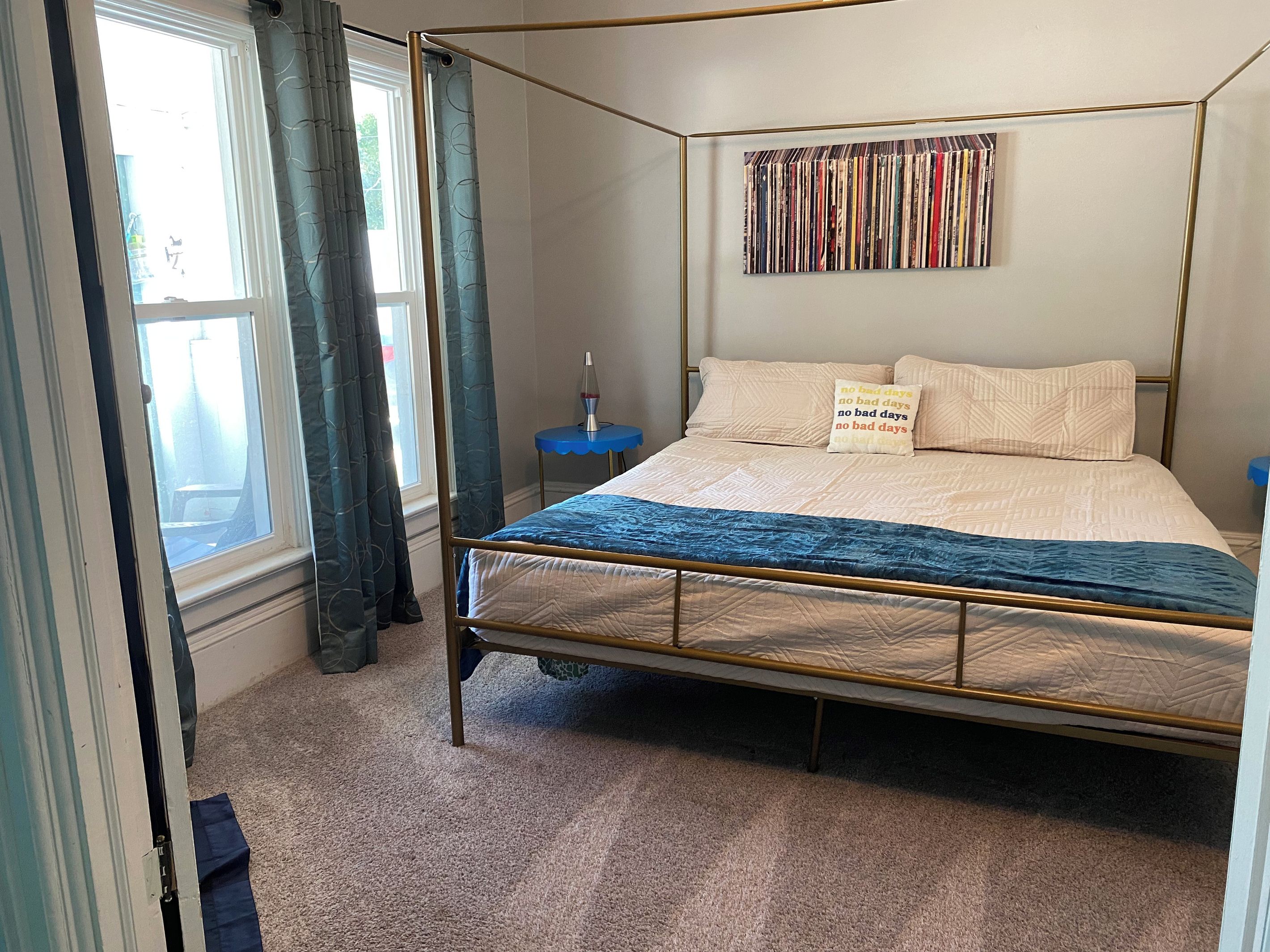 A four-poster bed with a blue bedspread, a wall-mounted bookcase, and a small blue side table with a lava lamp.