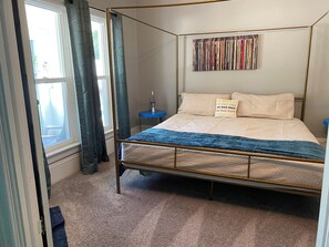 Ein Bett mit Baldachin, mit blauer Bettdecke, eine an der Wand befestigte Bücherreol und ein kleiner, blauer Nachttisch mit einer Lavalampe.