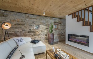 Una sala de estar acogedora con pared de piedra, un sofá blanco, una planta grande en maceta, una chimenea y una escalera de madera.