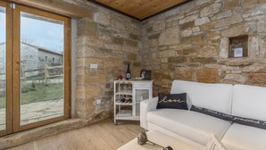 Una habitación acogedora con pared de piedra, un sofá blanco con cojín, un mueble pequeño con copas de vino y una ventana con vistas a un edificio.