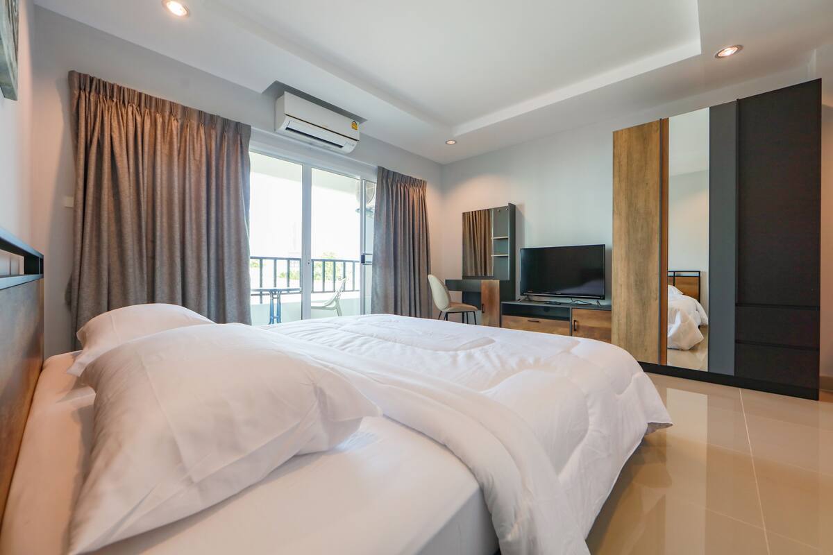 A hotel room with a large bed, a desk with a chair, a television, and a balcony with a view.