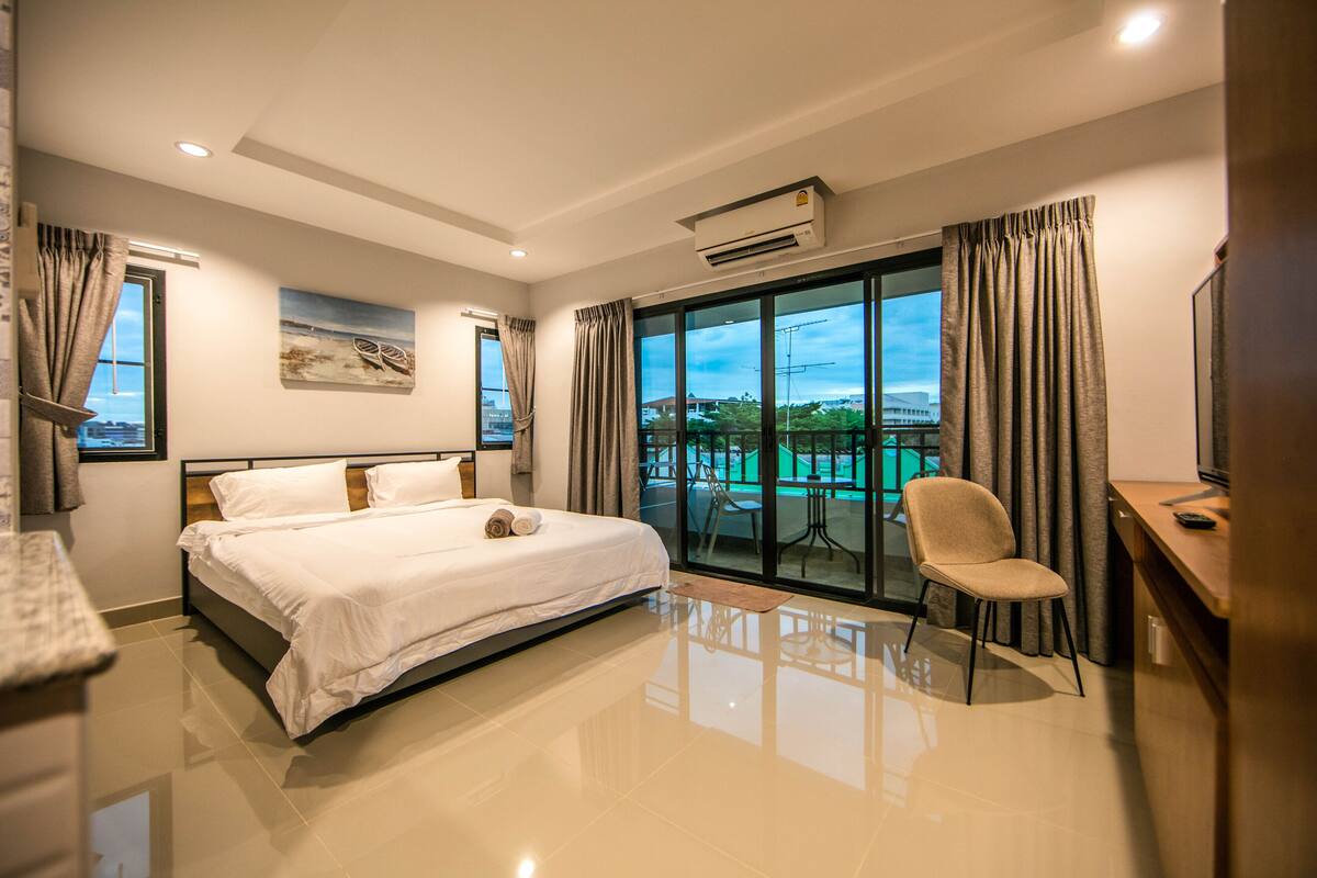 A modern hotel room with a large bed, a desk, and a chair. There is a balcony with a table and chairs outside the window.