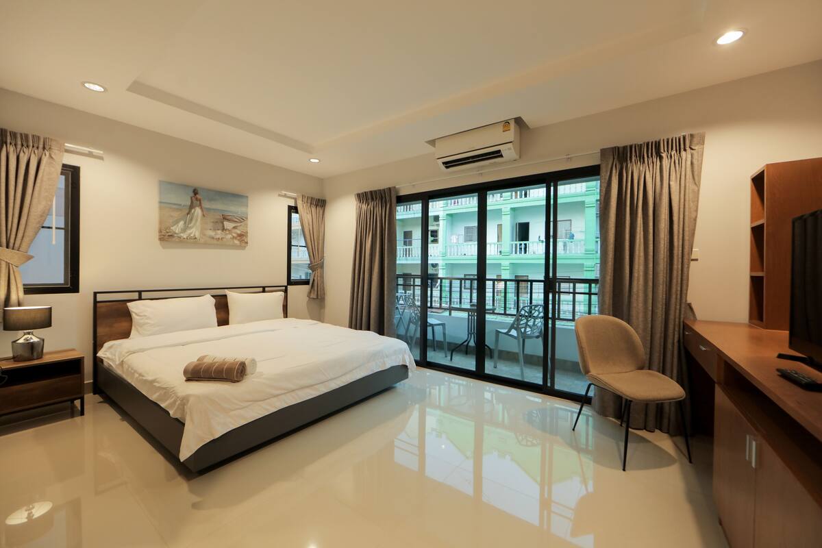 A modern hotel room with a large bed, a desk, a chair, and a balcony with a view of buildings.