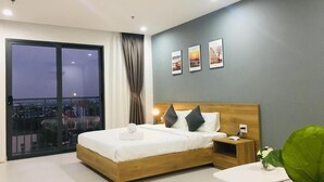 Una habitación de hotel moderna con una cama grande, una mesita de noche, una pequeña mesa y vistas a la ciudad a través de un ventanal amplio.