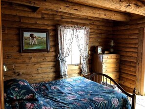 A wooden cabin room with a bed, a dresser, a framed picture on the wall, and a window with curtains. - Cliffside Log Cabin w/  Large Deck for Enjoying Spectacular Views & Wildlife (Boise)
