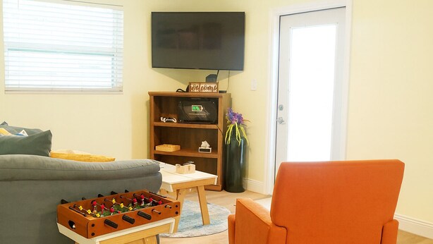 Un salón con un sofá, una mesa de centro de madera, una mesa de fútbol de mesa, un sillón naranja, un televisor montado en la pared y una estantería con un cartel de 'Welcome'.