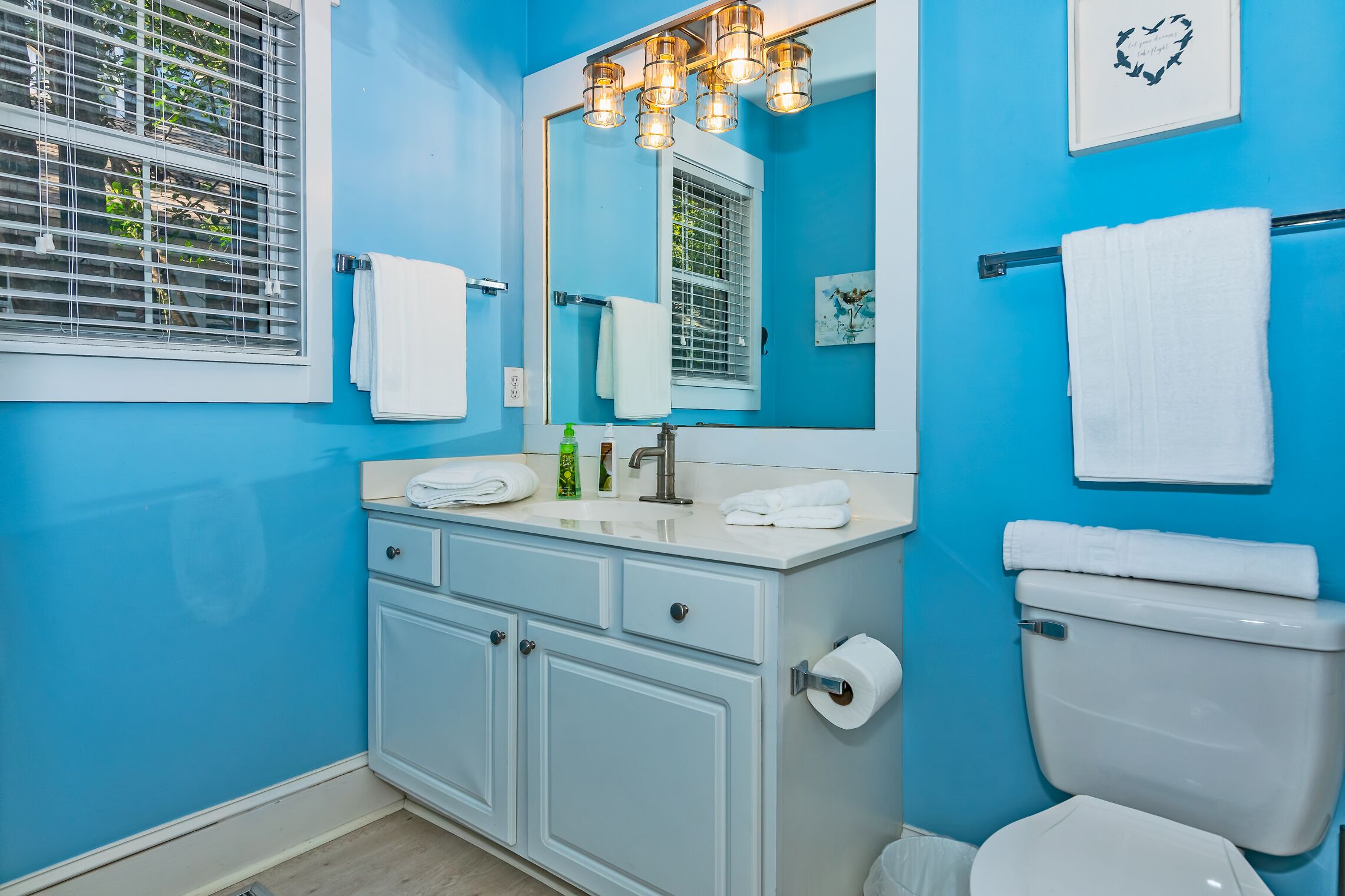 Un baño con pared azul, lavamanos blanco y inodoro. Hay toallas colgadas en la pared y una ventana con persianas.