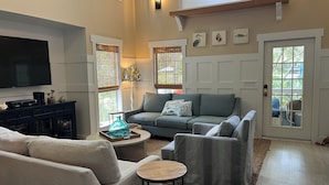 A spacious living room with a grey sofa, a coffee table, and a TV on a black cabinet. There are windows with blinds and a door with a view of the outside.