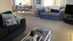 A living room with a dark blue sofa, a grey couch, and a coffee table with books and a lantern. There is a dining table with chairs and a blue bowl. - Pauanui Beach House. (Pauanui)