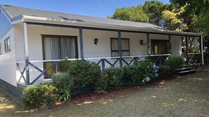 A single-story building with a porch, a satellite dish, and a small garden area with hydrangeas. - Pauanui Beach House. (Pauanui)