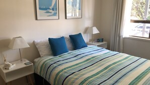 A neatly arranged hotel room with a bed, two nightstands, a lamp, a framed picture, and a window with curtains. - Pauanui Beach House. (Pauanui)