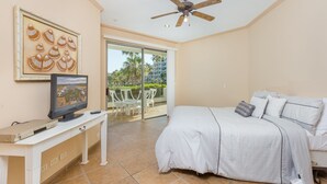 A bedroom with a bed, a TV, a ceiling fan, and a balcony with a view of palm trees. - Casa Blanca CB-102 Let Me Count The Waves Beautiful View Condo (Puerto Peñasco)