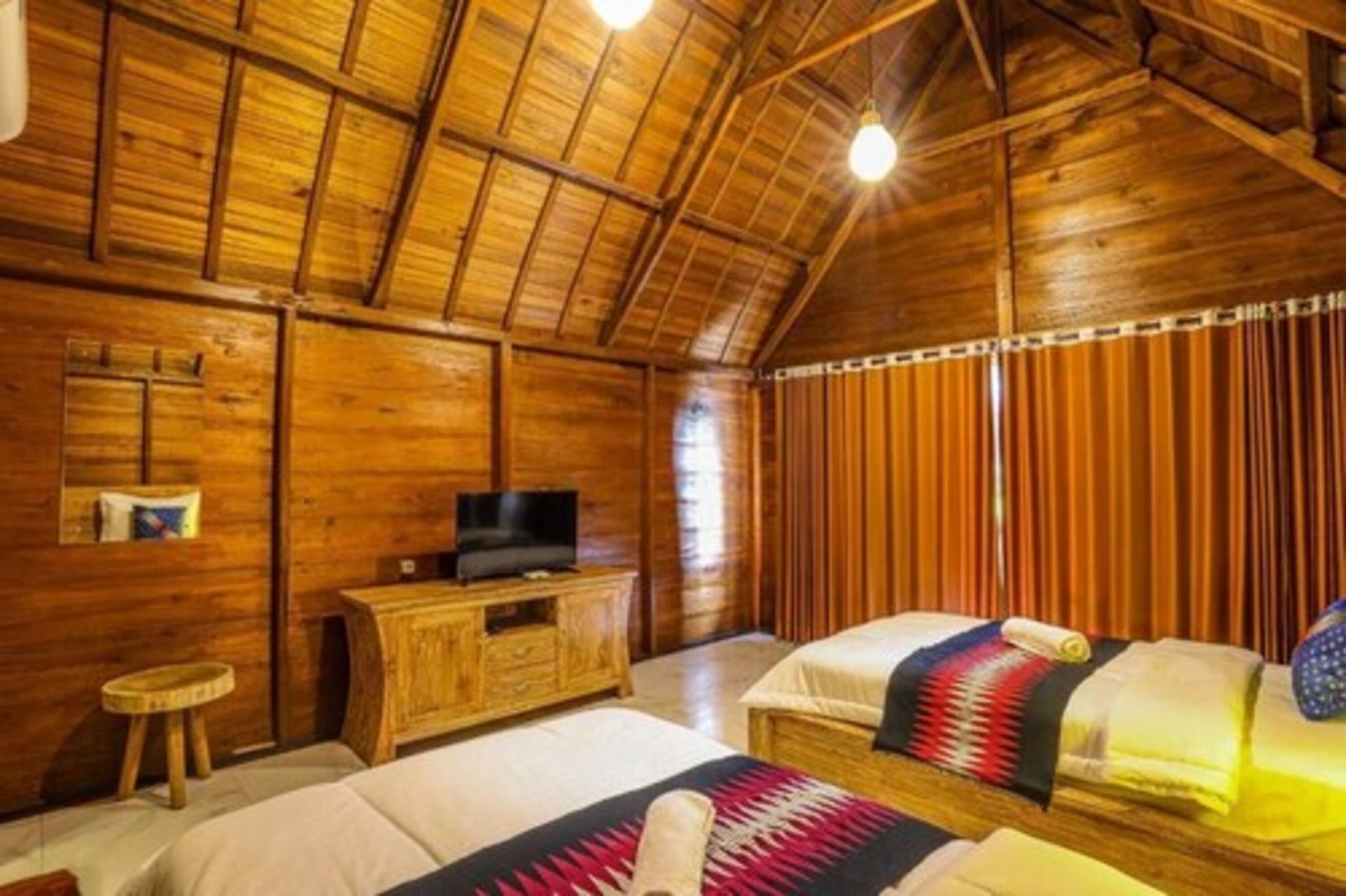 A wooden cabin-style room with two beds, a television on a wooden cabinet, and a window with vertical blinds.