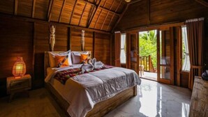 A wooden room with a bed, a nightstand, and a view of greenery outside. - The Tukad Gepuh Cottage (Penida Island)
