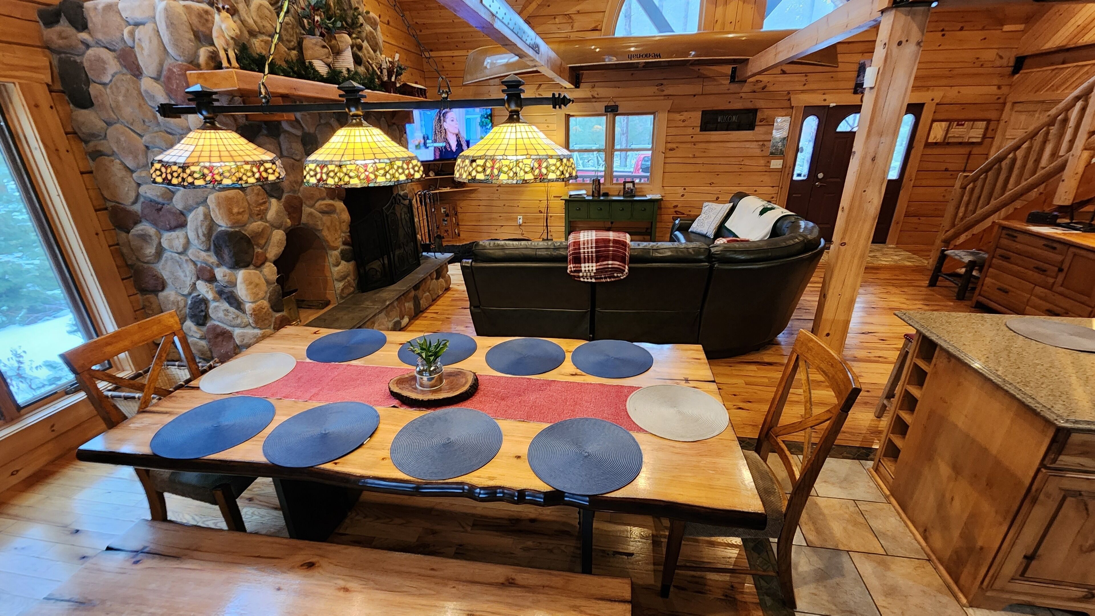 L’intérieur d’un chalet accueillant, avec une cheminée en pierre, une table à manger en bois agrémentée de sets de table, un canapé en cuir noir et un coin cuisine.