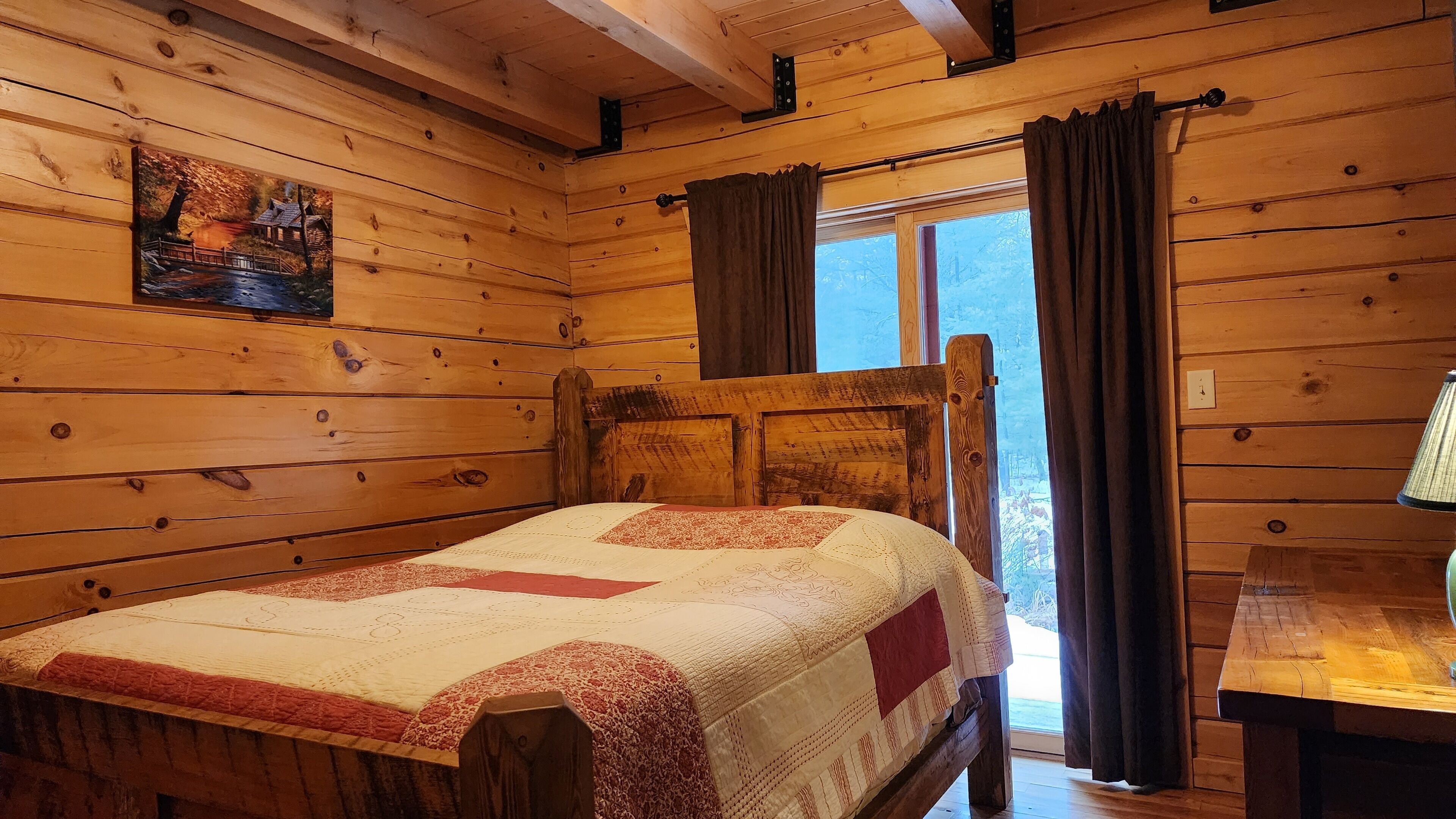 A wooden cabin bedroom with a bed, a painting, a window with curtains, and a lamp on a table.
