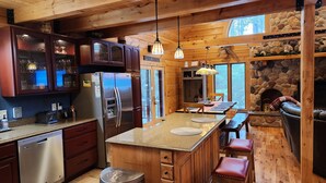 A rustic kitchen with wooden cabinets, a stainless steel refrigerator, a stone fireplace, and a dining area with a bench and table. - 🏞Secluded Log Cabin Near Wisconsin Dells & Wisconsin River • Fireplace • Ski 🏕 (Lyndon Station)