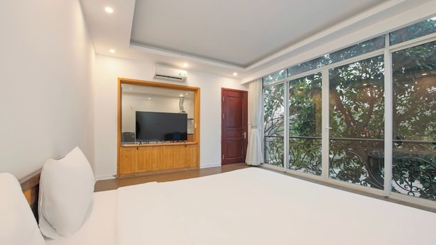 A bedroom with a large bed, a TV mounted on a wooden cabinet, a red door, and a balcony with a view of trees. - Lakeside House - Hanoi (Hanoi)