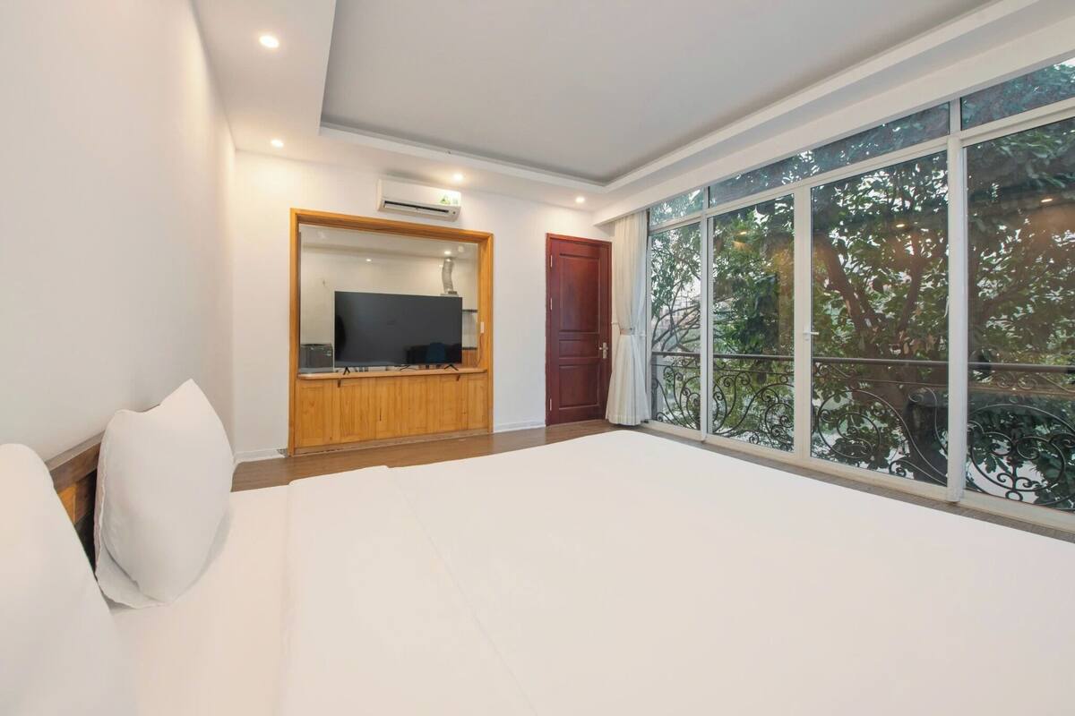 A bedroom with a large bed, a TV mounted on a wooden cabinet, a red door, and a balcony with a view of trees.