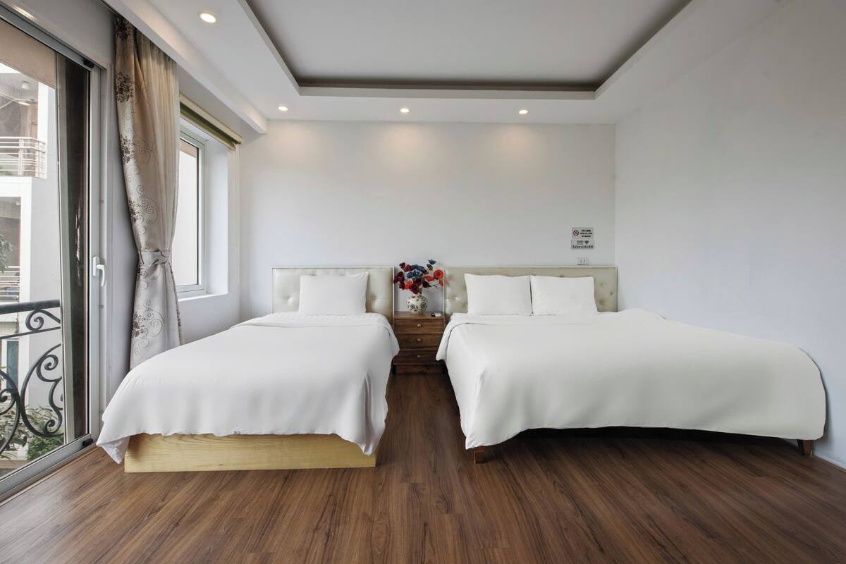 A hotel room with two beds, wooden flooring, a large window with curtains, and a small bedside table with a vase of flowers.