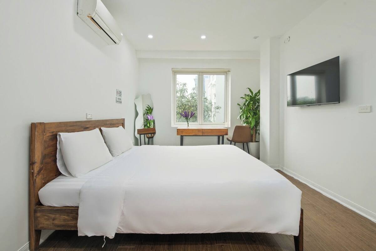 A bedroom with a wooden bed, a desk, a chair, a TV, and a window with a view of greenery.