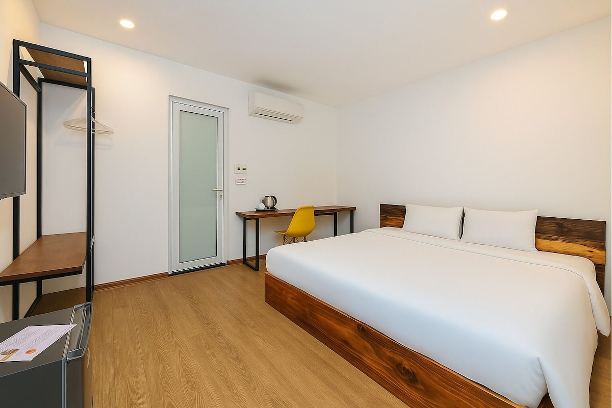 A hotel room with a bed, a desk, a chair, a wardrobe, and an air conditioning unit.