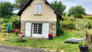 A small house with a stone foundation, a pitched roof, and a single window with shutters. There is a small garden area with a table and chairs, and a satellite dish on the roof.