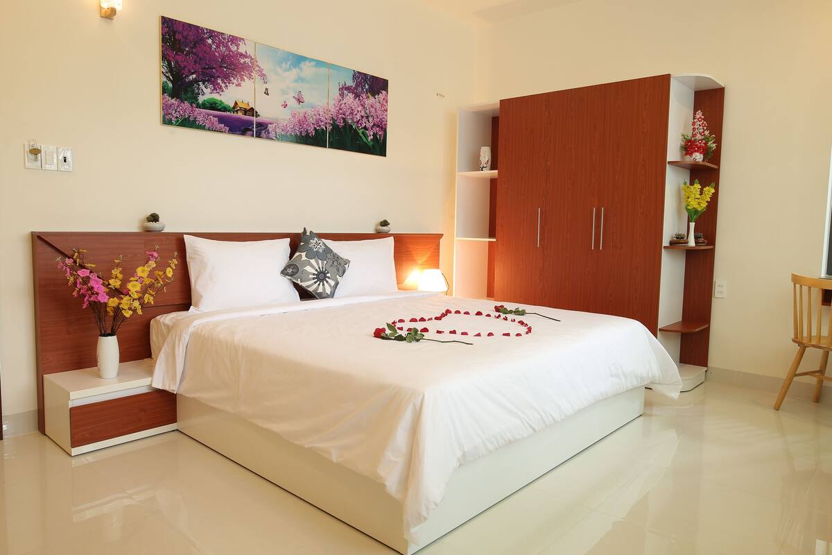 Een netjes opgemaakt bed met witte beddengoed en een rozenboeket, een houten garderobe, een nachtkastje met een bloevas en een bloemen schilderij aan de muur.