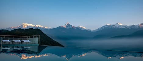 A infinity pool reflecting mountains, a modern building with lounge chairs, and a clear sky.
