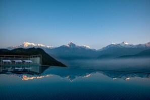 A infinity pool reflecting mountains, a modern building with lounge chairs, and a clear sky.