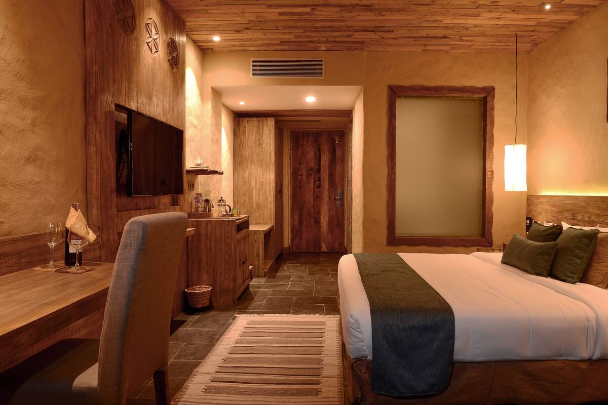 A hotel room with a wooden headboard, a bed with white linens, a desk with a chair, a small table with a wine glass, and a wall-mounted TV.