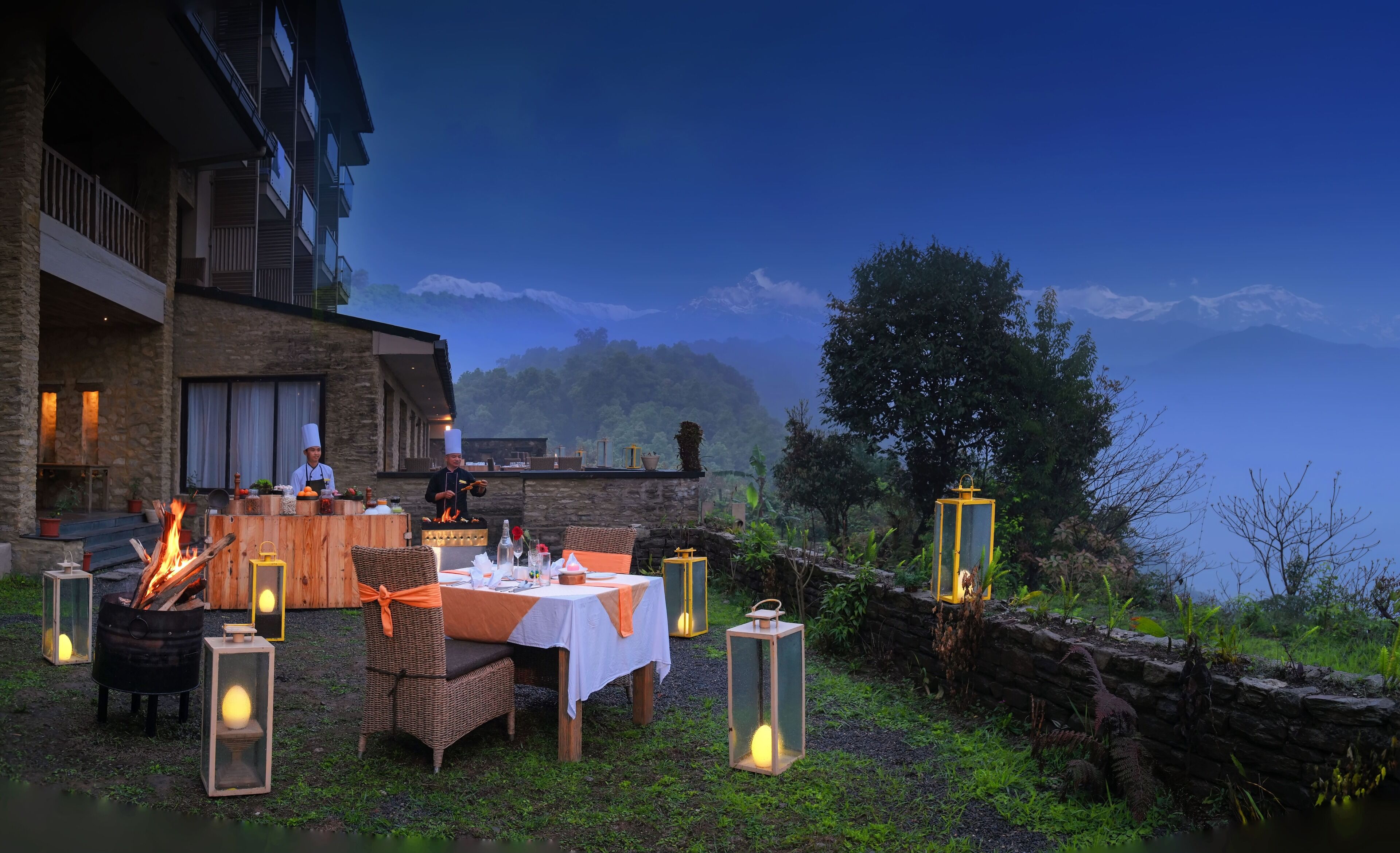 An outdoor dining area with a stone building, a wooden table set for two, and a fire pit.