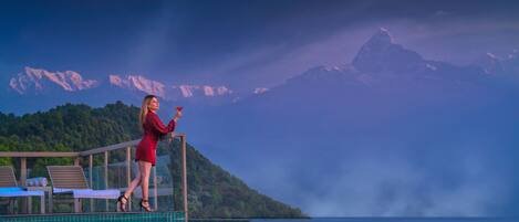 A woman in a red dress standing by a glass railing overlooking a pool with a mountain backdrop.