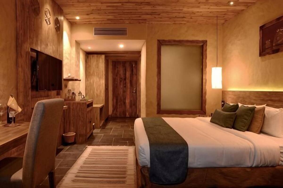 A hotel room with a wooden ceiling, a bed with pillows, a desk with a chair, a television, and a bathroom door.