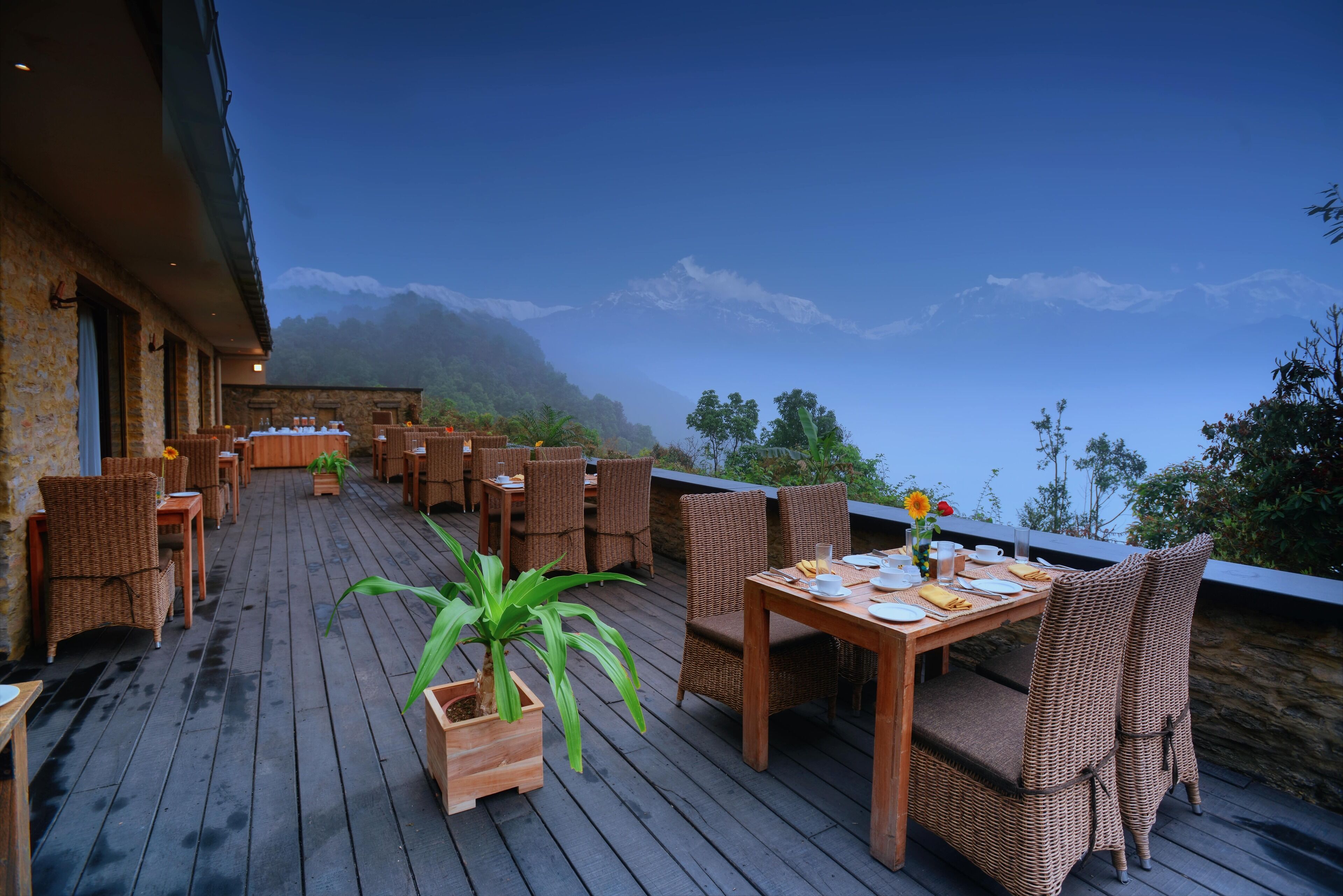 An outdoor dining area with wicker furniture and a table set for two, overlooking a mountainous landscape.