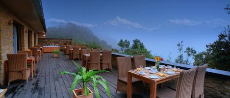 An outdoor dining area with wicker furniture and a table set for two, overlooking a mountainous landscape.