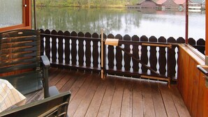 A wooden deck with a wooden railing, a bench, and a view of a lake with buildings in the background. - Bootshaus in Mirow mit überdachter Terrasse (Mirow)