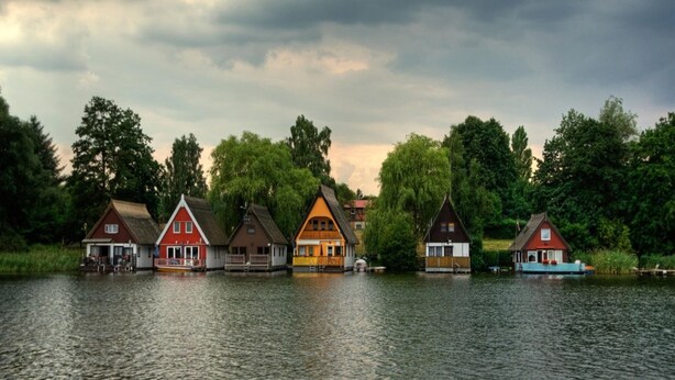A row of traditional wooden houses by a lake with a cloudy sky. - Bootshaus in Mirow mit überdachter Terrasse (Mirow)