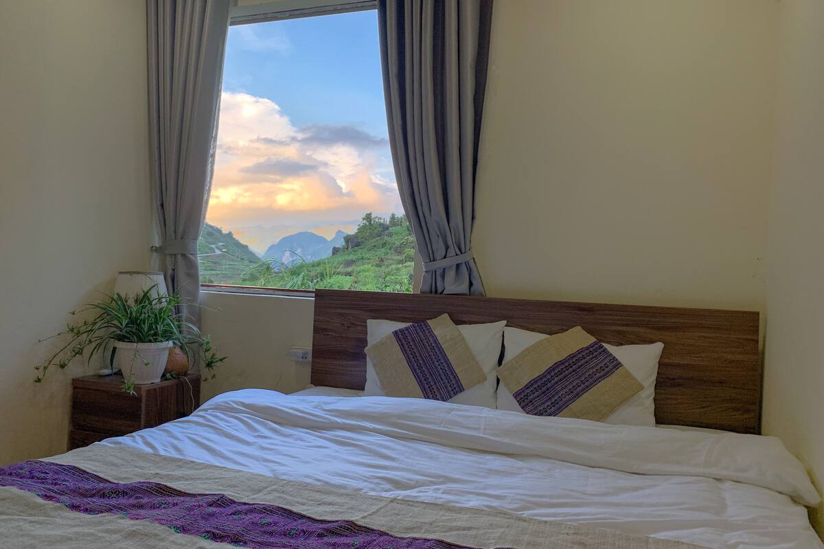 A bedroom with a bed, a window with curtains, a nightstand with a plant, and a view of mountains.