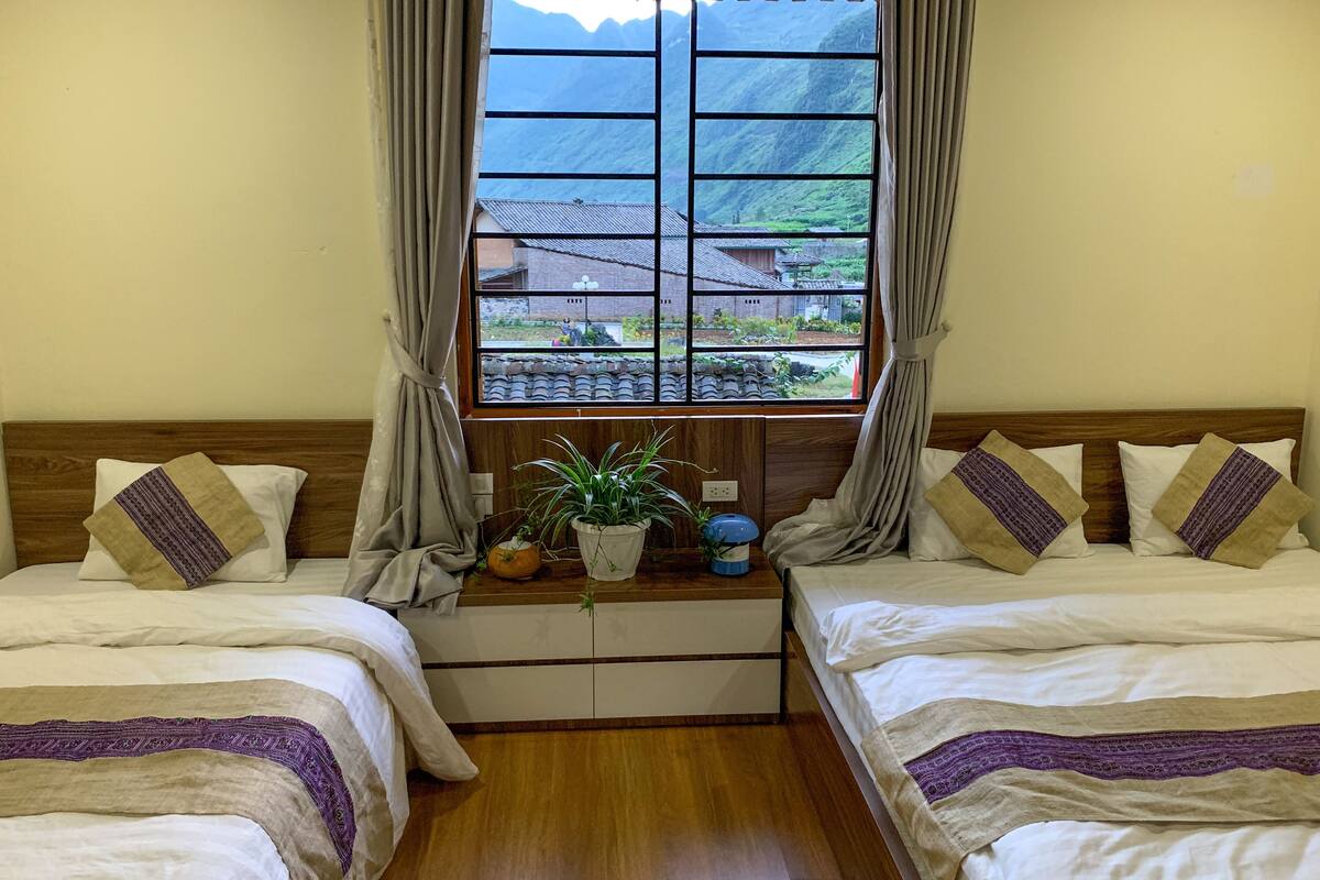 A room with two beds, a window with a view of buildings and mountains, and a small table with a plant.