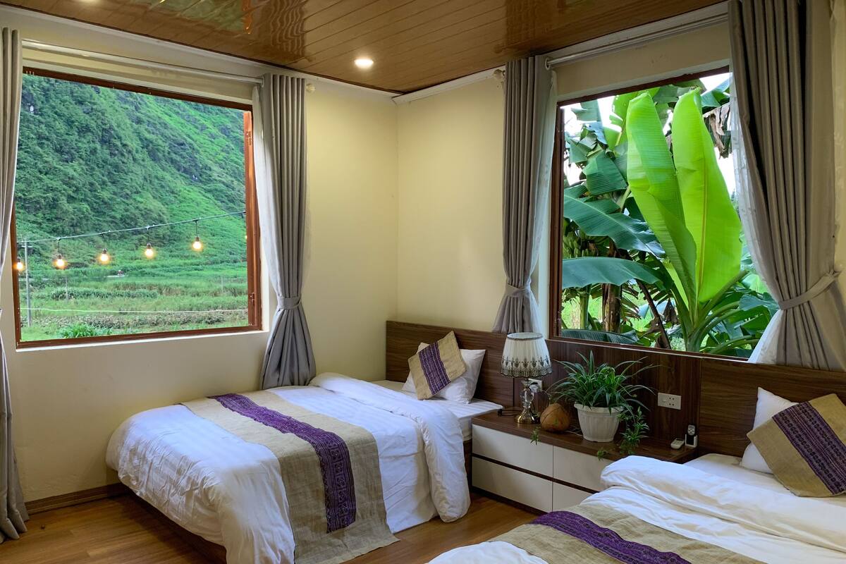 A bedroom with two beds, a nightstand, a lamp, and a view of a lush green landscape through large windows.
